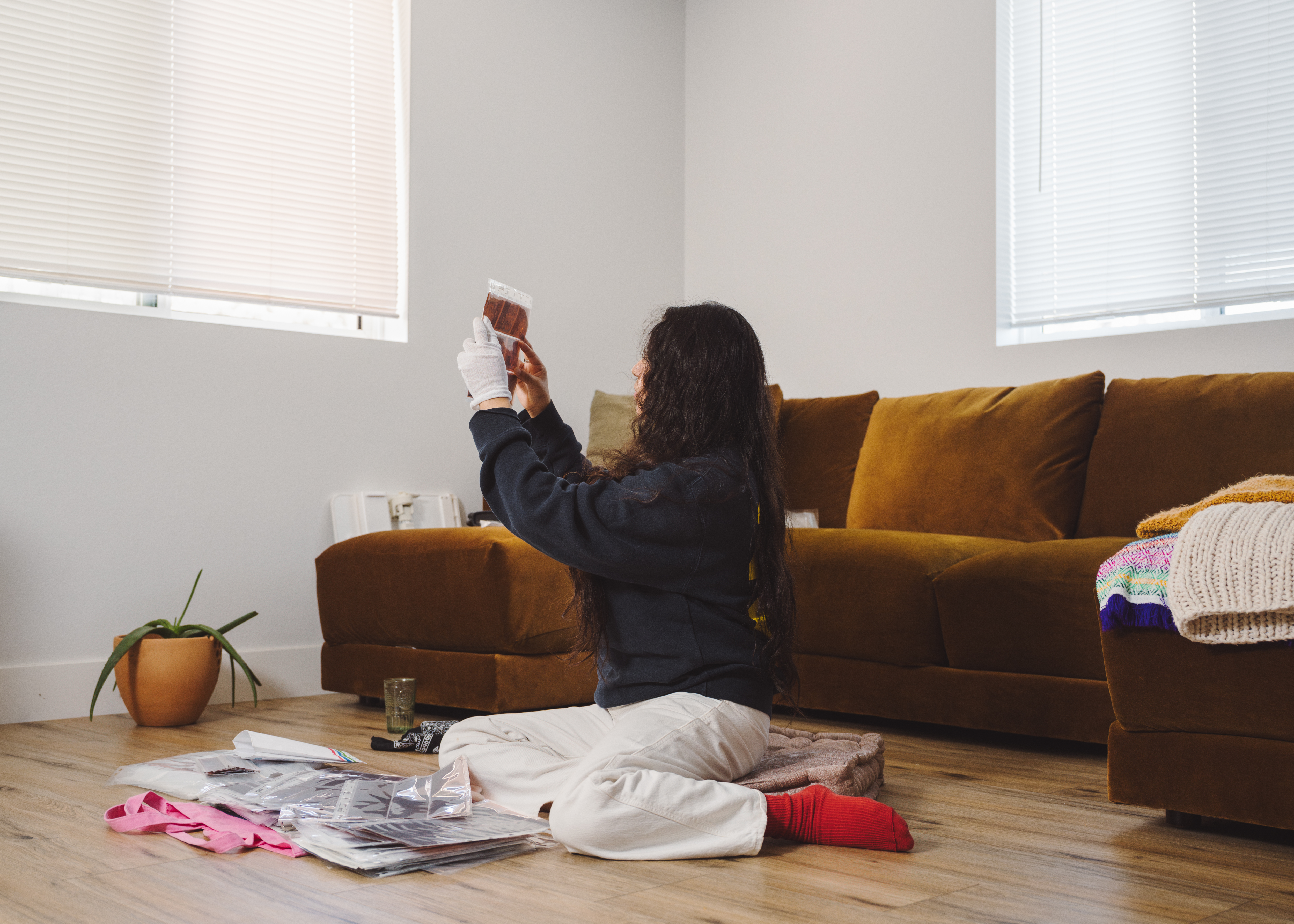 A young woman, facing sideways away from the camera, sits on the living room floor, holding photo negatives up toward a window.
