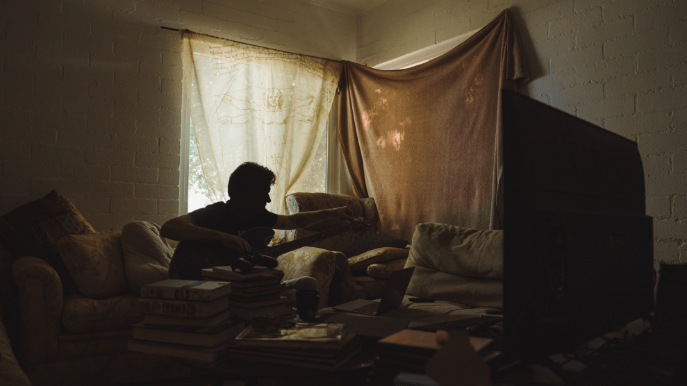 A person in a dimly lit room, sitting on a couch while tuning his guitar.