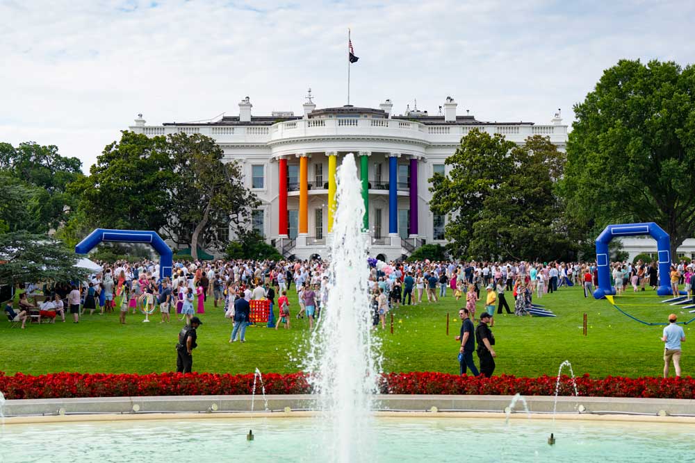 Hundreds of people fill the White House lawn. The six pillars of the semicircular portico facing the South Lawn are wrapped in the rainbow colors of the Pride flag.