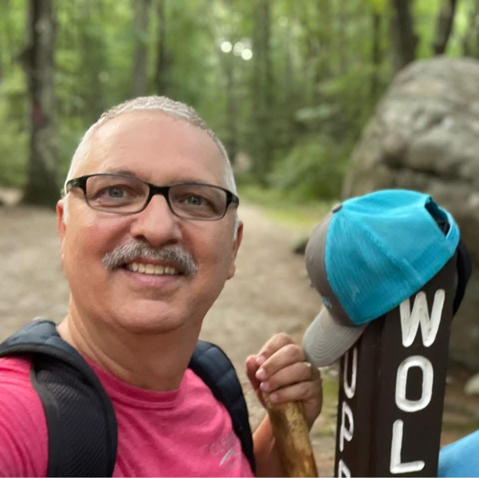 A smiling middle-aged James Harter with gray mustache, glasses, and short gray hair poses for a photo in the wilderness. He holds a walking stick and his gray-and-blue baseball hat rests on a nearby trail marker post.
