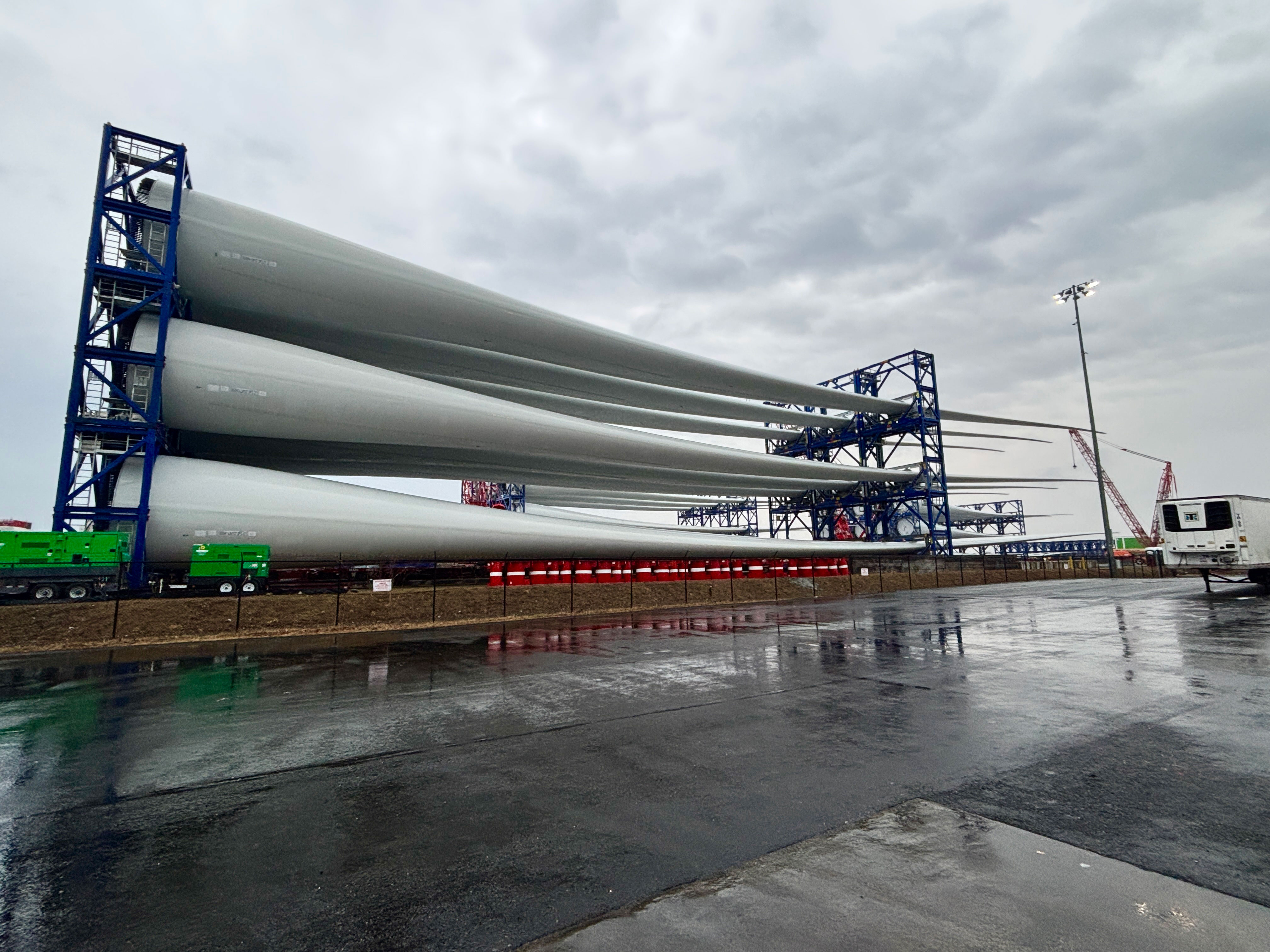 Giant white turbine blades sit on a massive horizontal rack on a cloudy day in New England