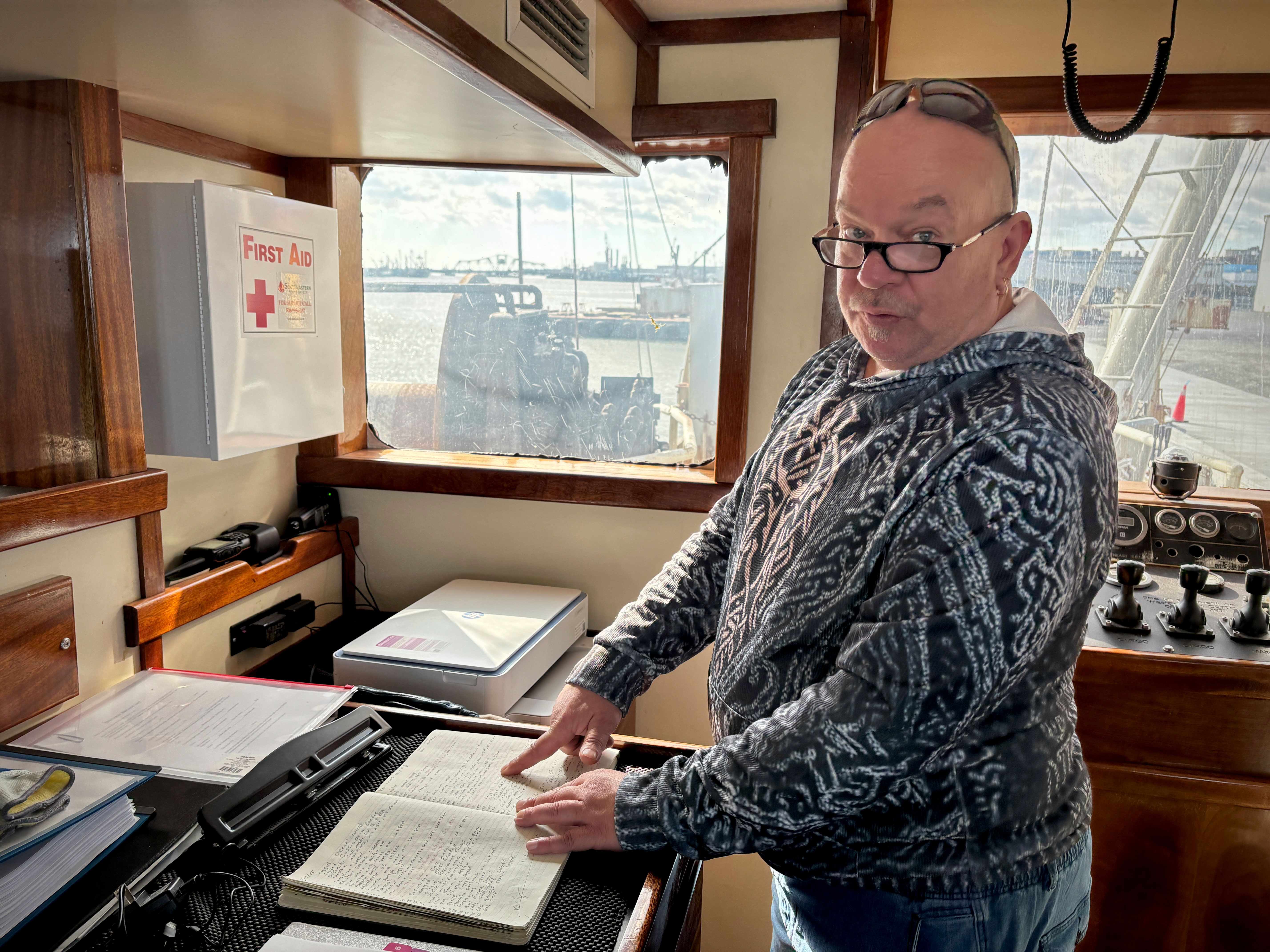 A portly white man with a shaved head, reading glasses, and a blue sweater in a ship's cabin, is pointing to a page in an open book while looking up to the side at the photographer.