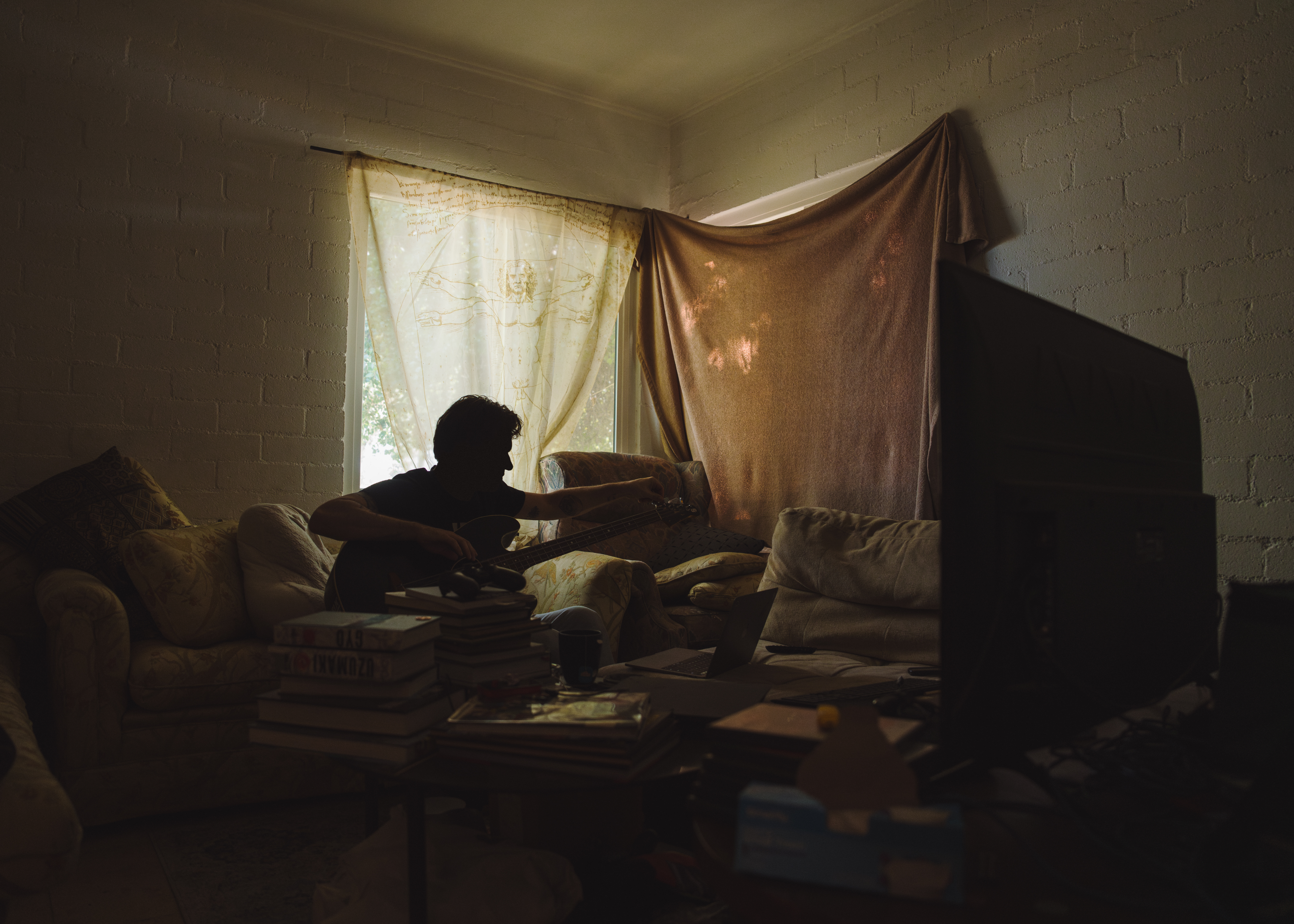 A man, backlit against a window in a dimly lit room, sits on a couch while tuning a guitar.