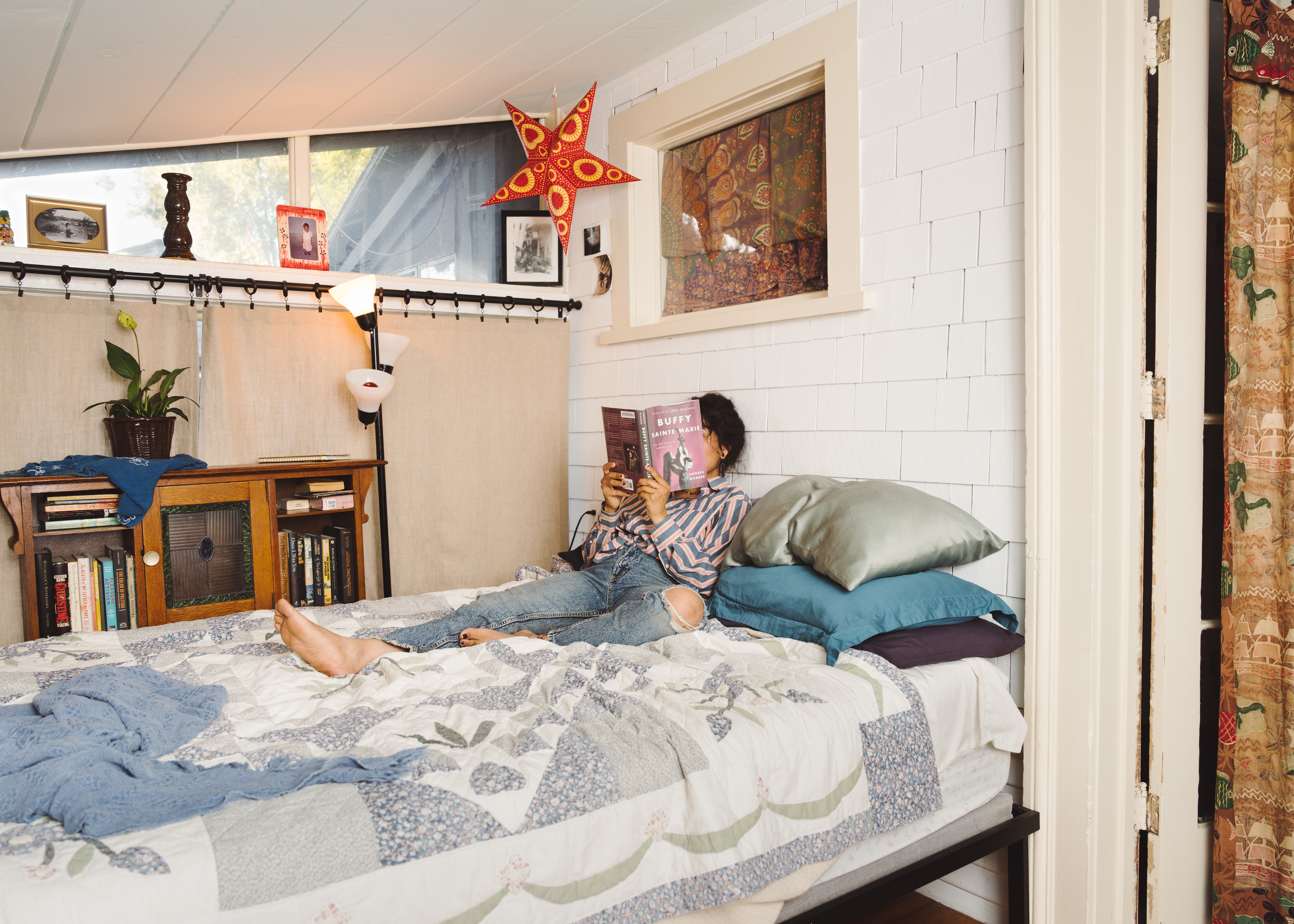 A woman sits on a quilt-covered bed, holding a book that covers her face.
