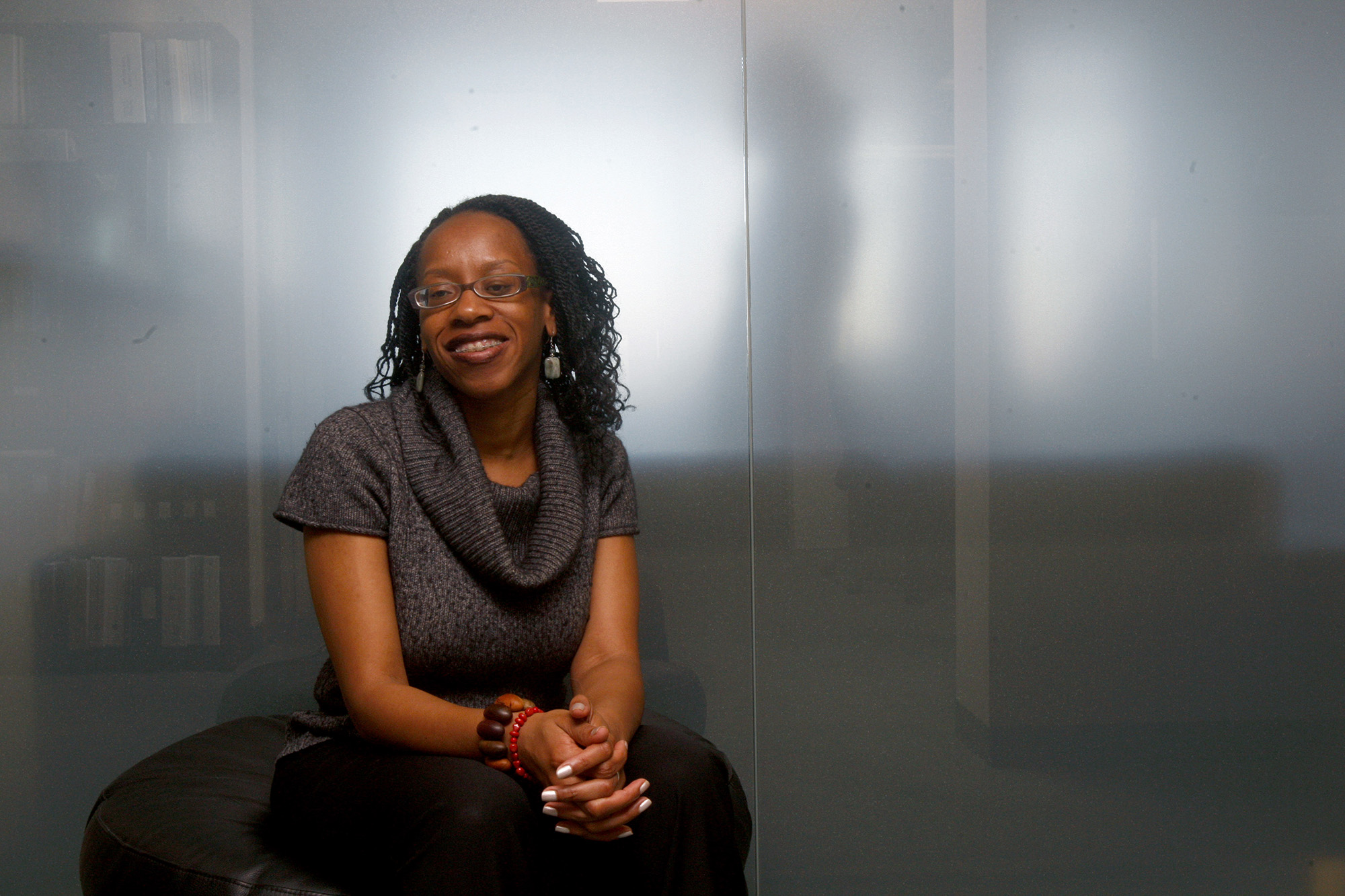 Portrait of Lateefah Simon, youngish black woman, seated and smiling with a large neck turtleneck short-sleeved sweater and a large bracelet with wooden beats. Background is clouded glass windows of an office or conference room.