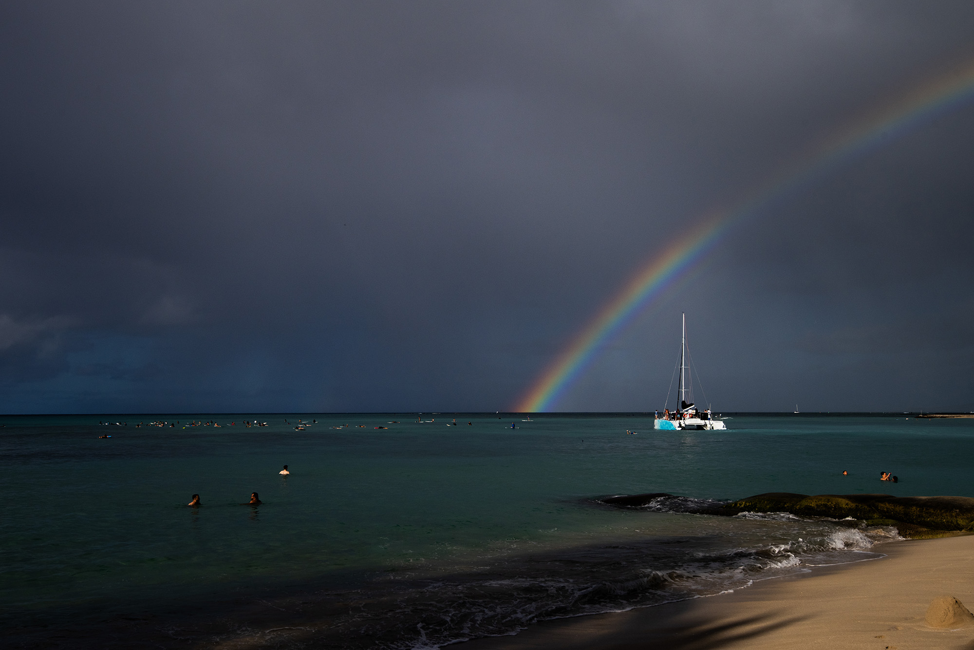 Rainbow over the water with cloudy skies and a catamaran, sails down, underneath, and some people swimming in the water near a beach. 