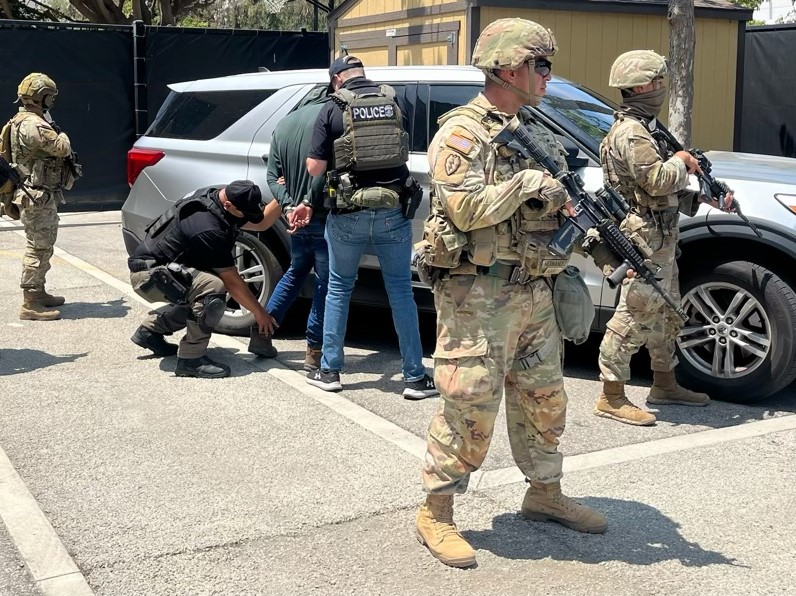 Three armed National Guard members stand around a silver SUV while two law enforcement officers search and detain a man
