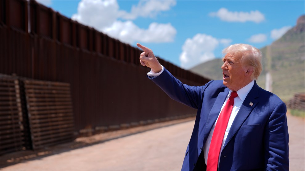 Donald Trump points to the southern border with his mouth open. He is wearing a navy blue suit, a red tie, and an American flag pin.
