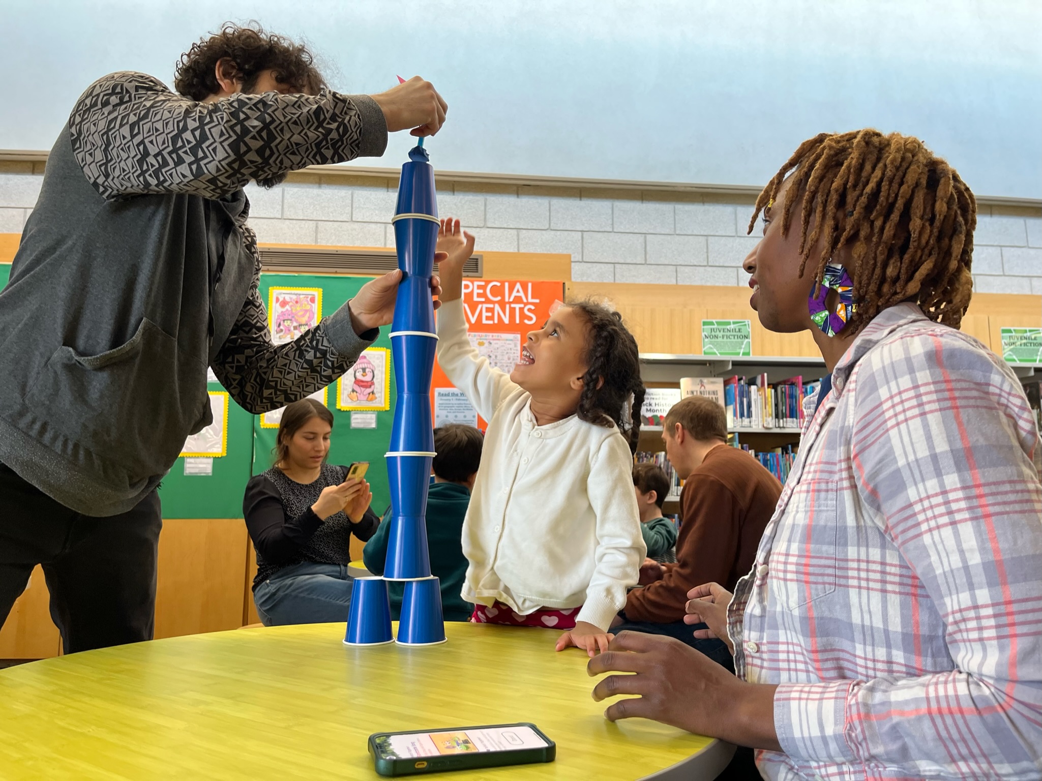 A a smiling child stacking blue cups with two adults at a table.