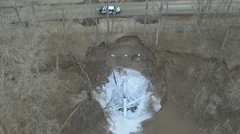 Aerial shot of a road with a single car, trees with no leaves, and brown earth. At the center is a crater in the ground, coated in ice with a pipe in the center.