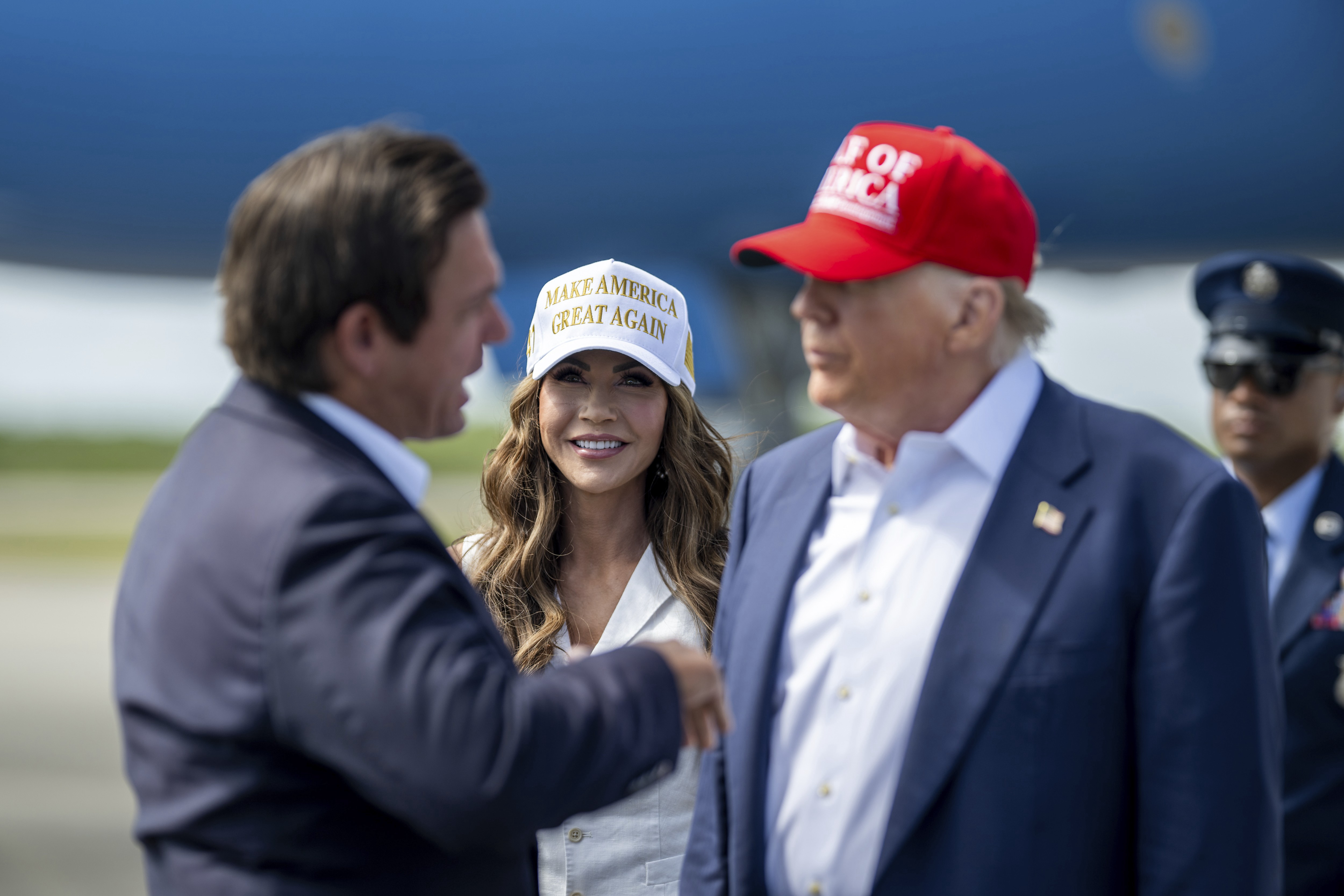 Kristi Noem, wearing a white MAGA baseball hat, smiles as Ron DeSantis approaches Donald Trump for a handshake. 