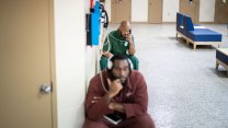 In a sparse institutional space with some blue furniture and multiple cell doors, two black men, one in blue and one in red clothing, face the camera as the speak on telephones lined along one wall.