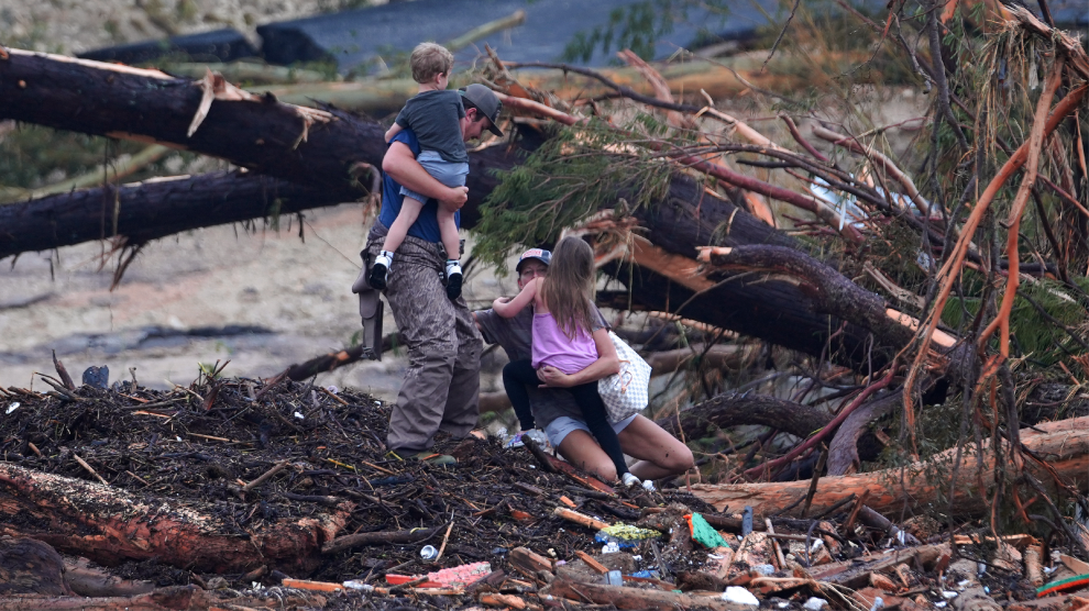 A woman in shorts and a baseball cap balances on her knees atop a pile of fallen trees and other debris while holding a little girl. A man holding a little boy stands beside her.