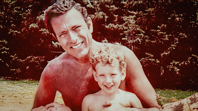 Smiling, care-chested white father and son in the early 1970s, color photo, background is green conifer shrubs