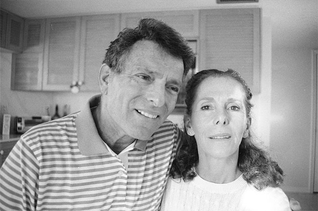 An older Harvey Schein and his wife Joy in the kitchen at their place in Florida. Black and white photo. Harvey wears a striped Rugby style shirt. joy wears a white t-shirt.