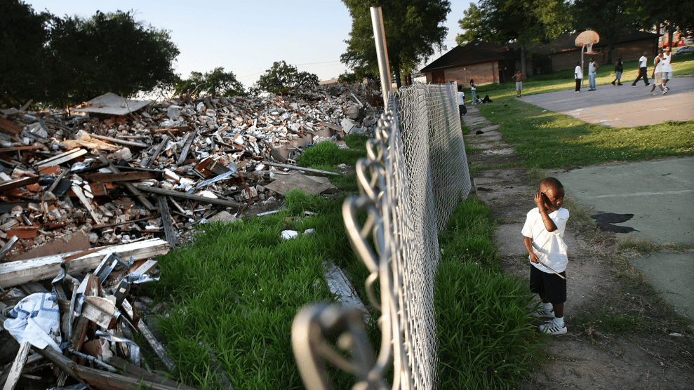 A photo divided by a chainlink fence. On the right we see people playing basketball on a blacktop in the background. In the foreground is a child in a white t-shirt and shorts looking back over his shoulder. On the other side of the photo, and the fence, is a massive pile of debris.