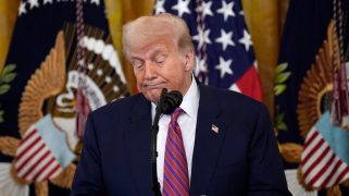 President Donald Trump standing in front of three flags and behind a microphone, sheepishly looking downwards.