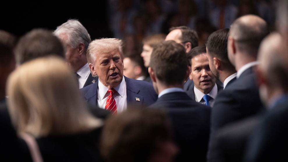 Donald Trump, wearing a blue suit and red tie, stands in the middle of a crowd and speaks to someone off-camera. Secretary of State Marco Rubio is visible over Trump’s left shoulder.