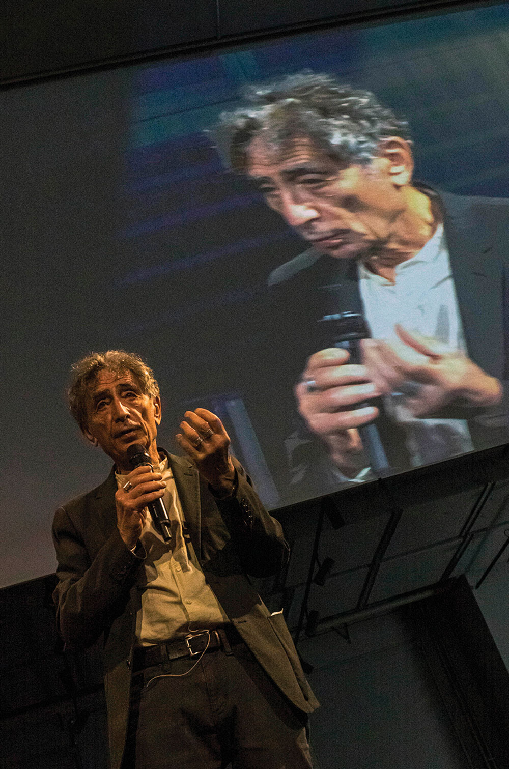 An elderly man wearing a suit without a tie with gray, curly hair extends his left hand with curled fingers, holding a microphone with his right hand. His public talk is being displayed on a screen behind him.