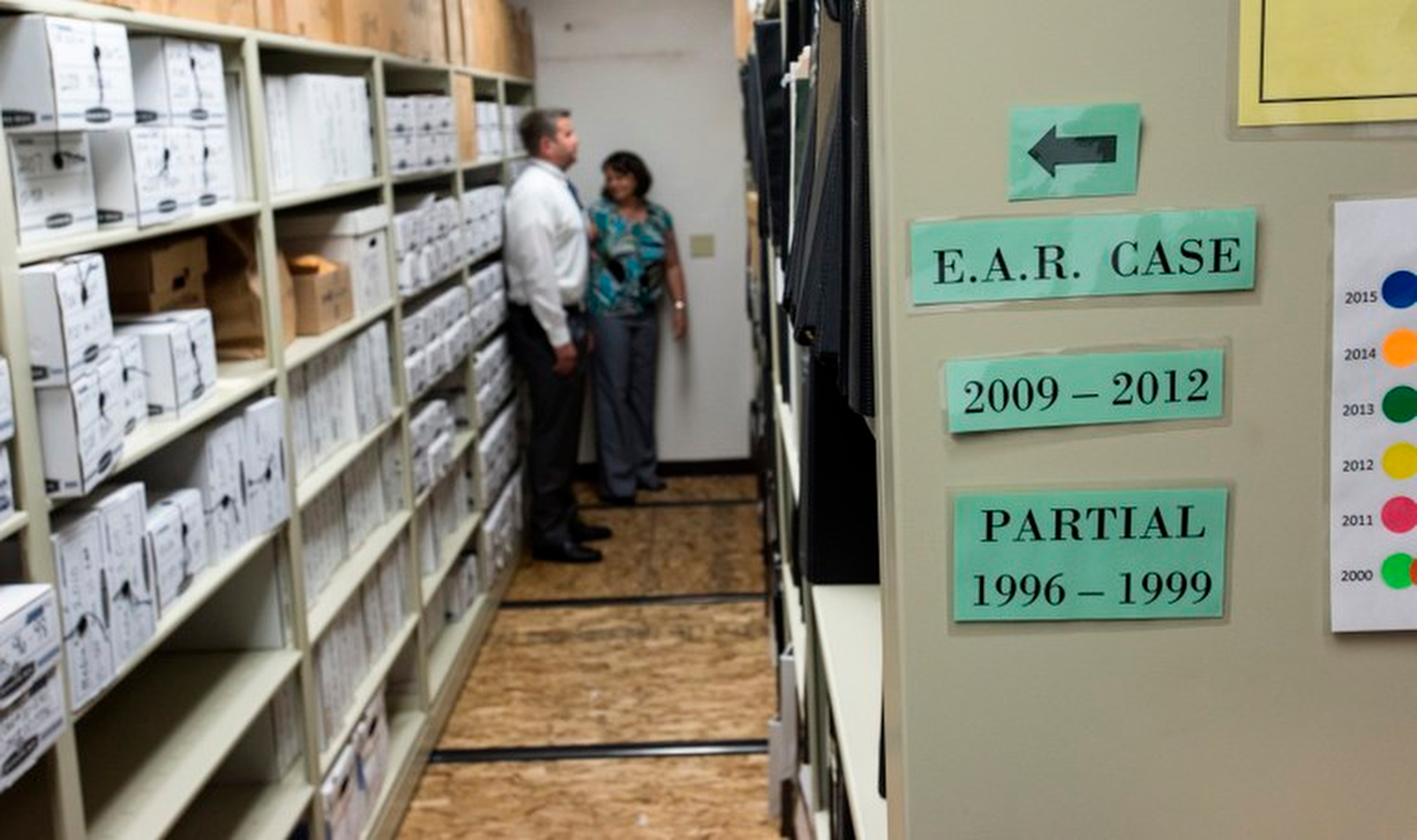 Two people stand at the end of an aisle lined with metal shelving units, filled with boxes.