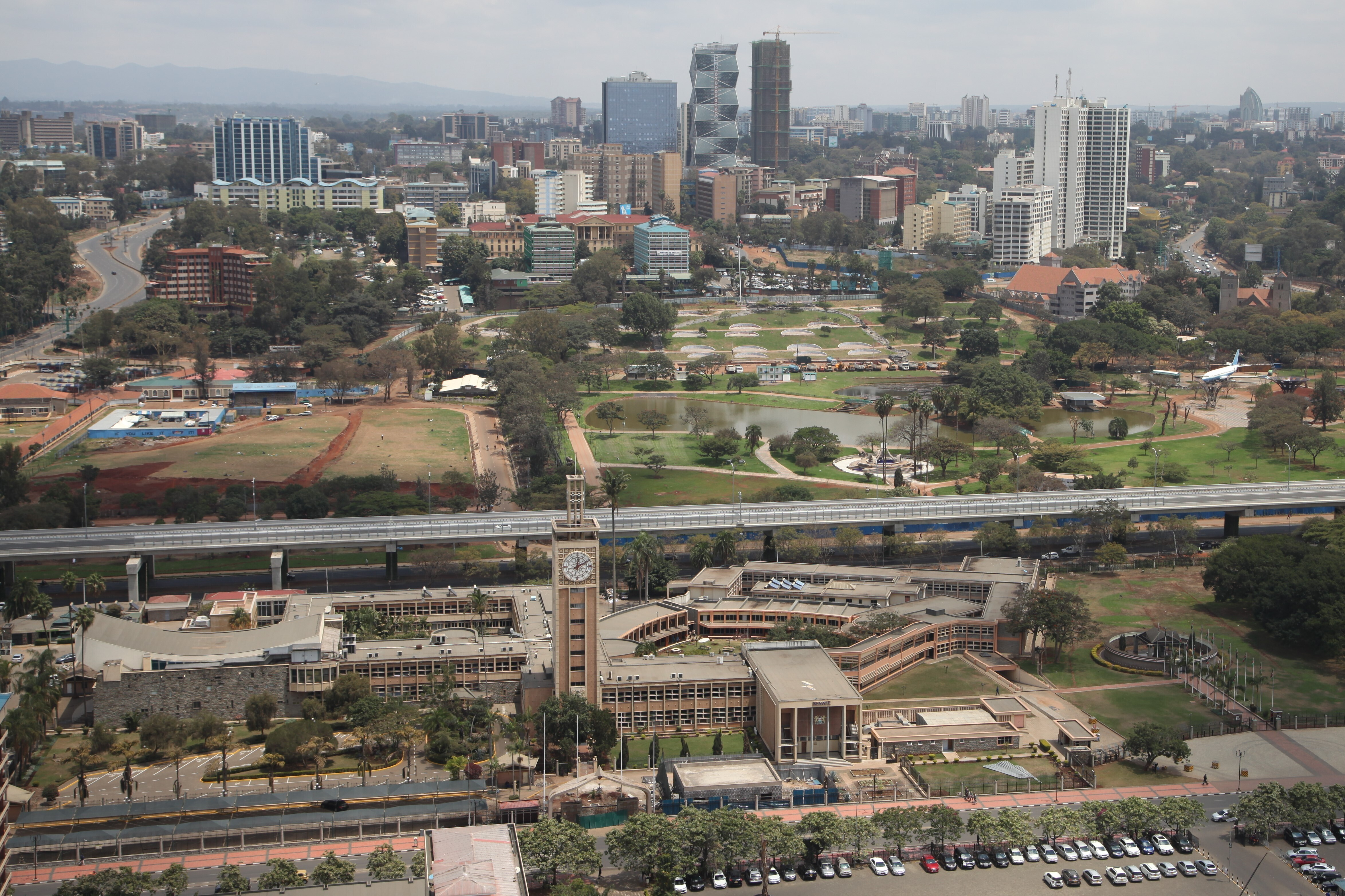 An aerial photo of the modern Kenya Parliament building, with a tall clock tower. Beyond it are green fields of a park and tall downtown buildings.