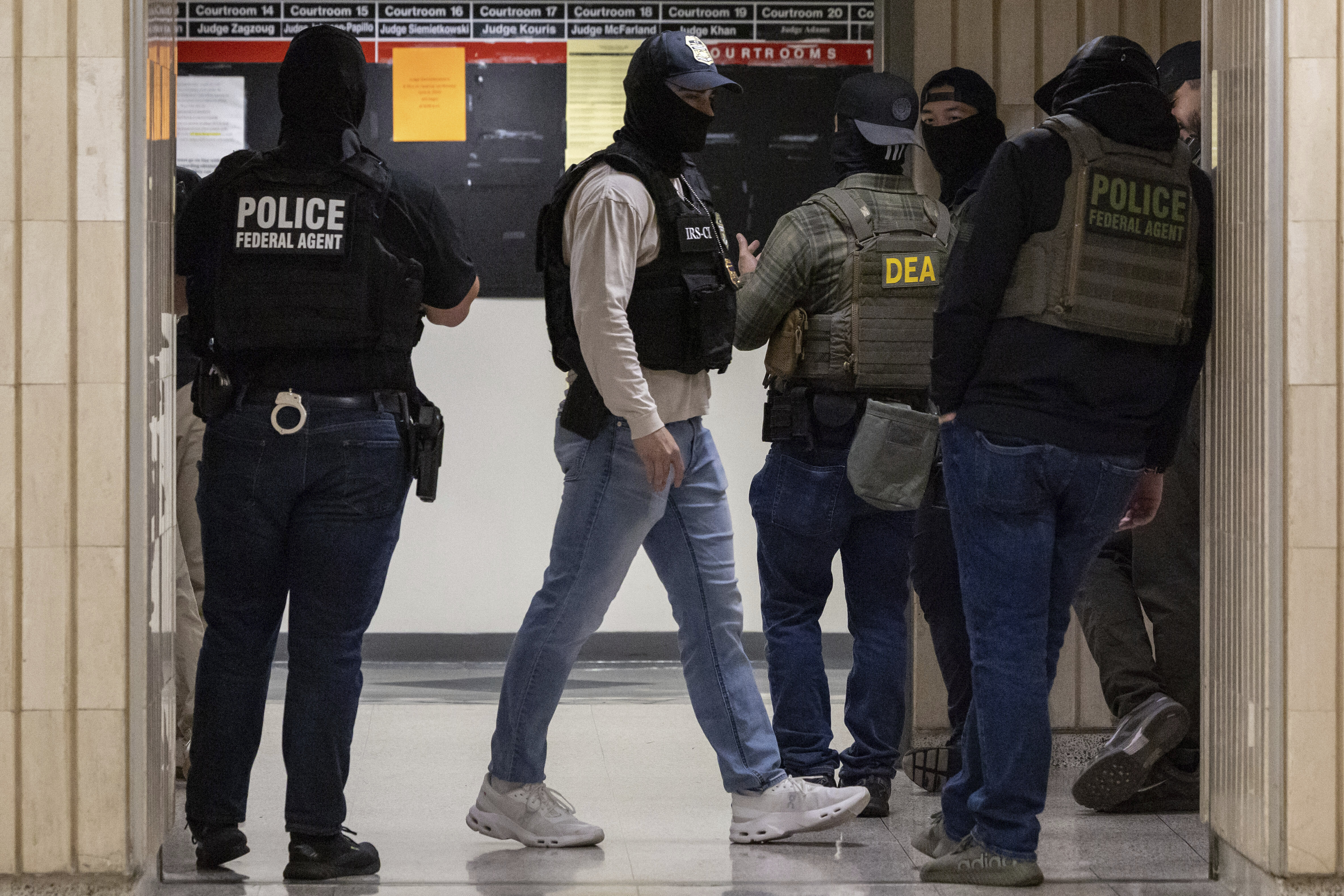 Federal agents walk around a hallway in jeans, baseball caps, sneakers, facial coverings, and tactical vests. 
