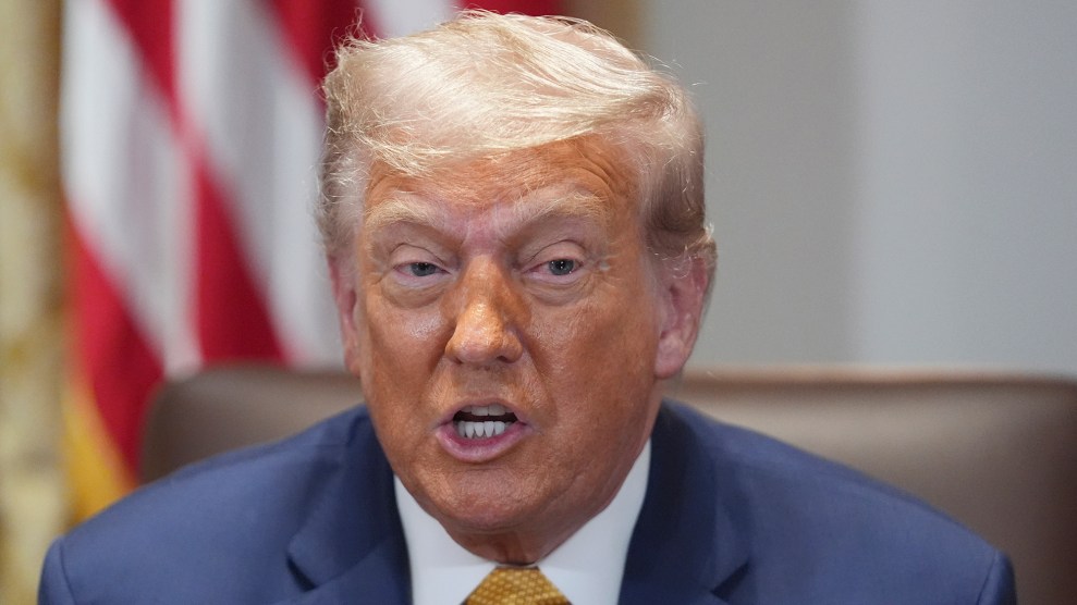 Donald Trump wears a blue suit and sits in front of an American flag. His mouth is partially open.