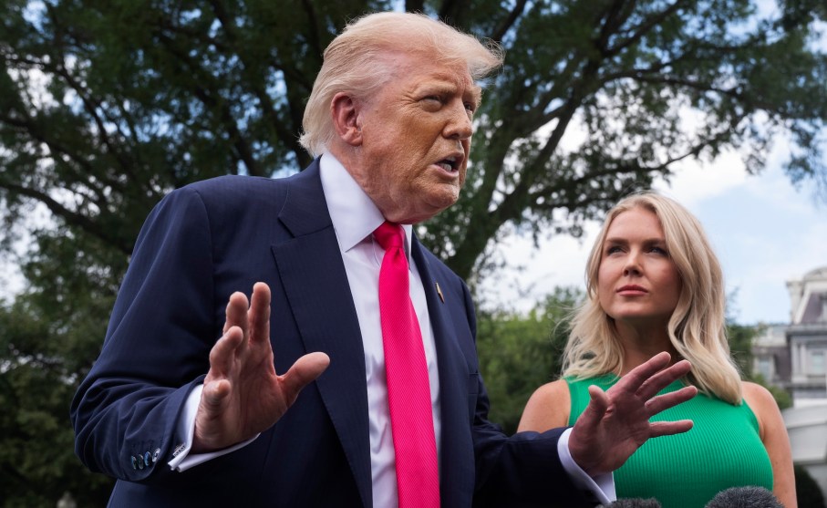 President Donald Trump in a suit and red tie gestures with his hands while speaking outside under a leafy tree. White House press secretary Karoline Leavitt stands beside him, slightly out of focus, looking at him.