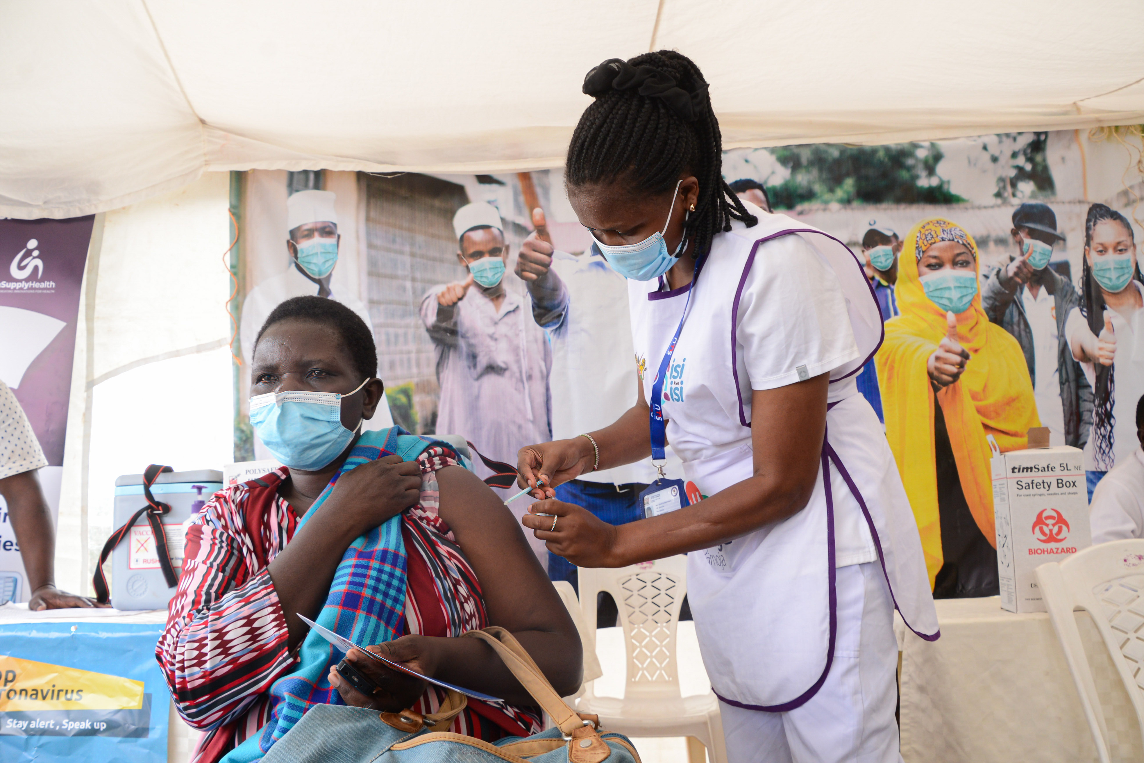A health care worker injects a shot into a woman's upper arm. Both are wearing blue paper medical face masks.