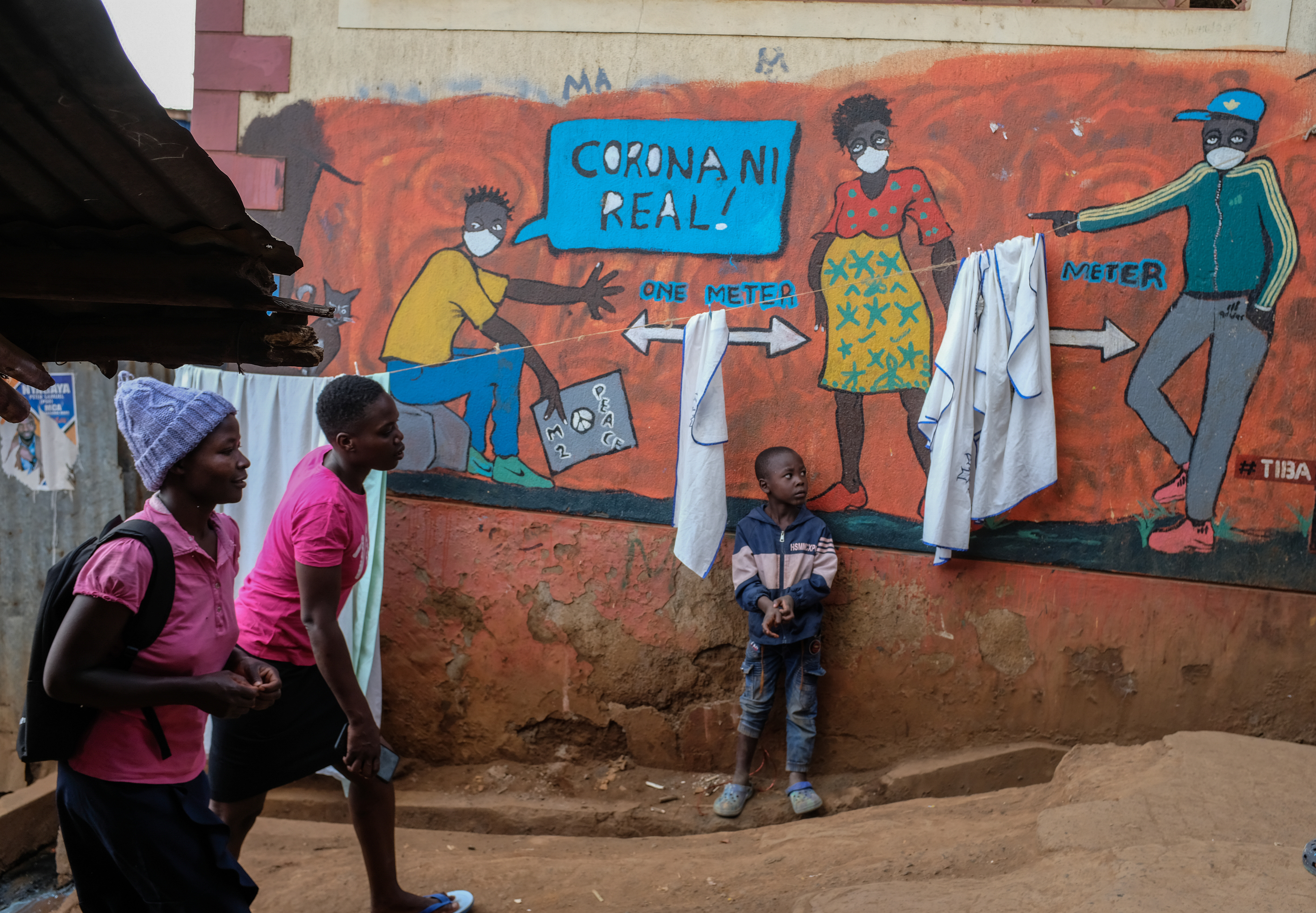 A young Kenyan boy stands in front of a brightly painted mural. In the mural, three people wear white face masks, and arrows indicate a distance of 1 meter between the people. One person in the mural says in Swahili, "Corona is real!"