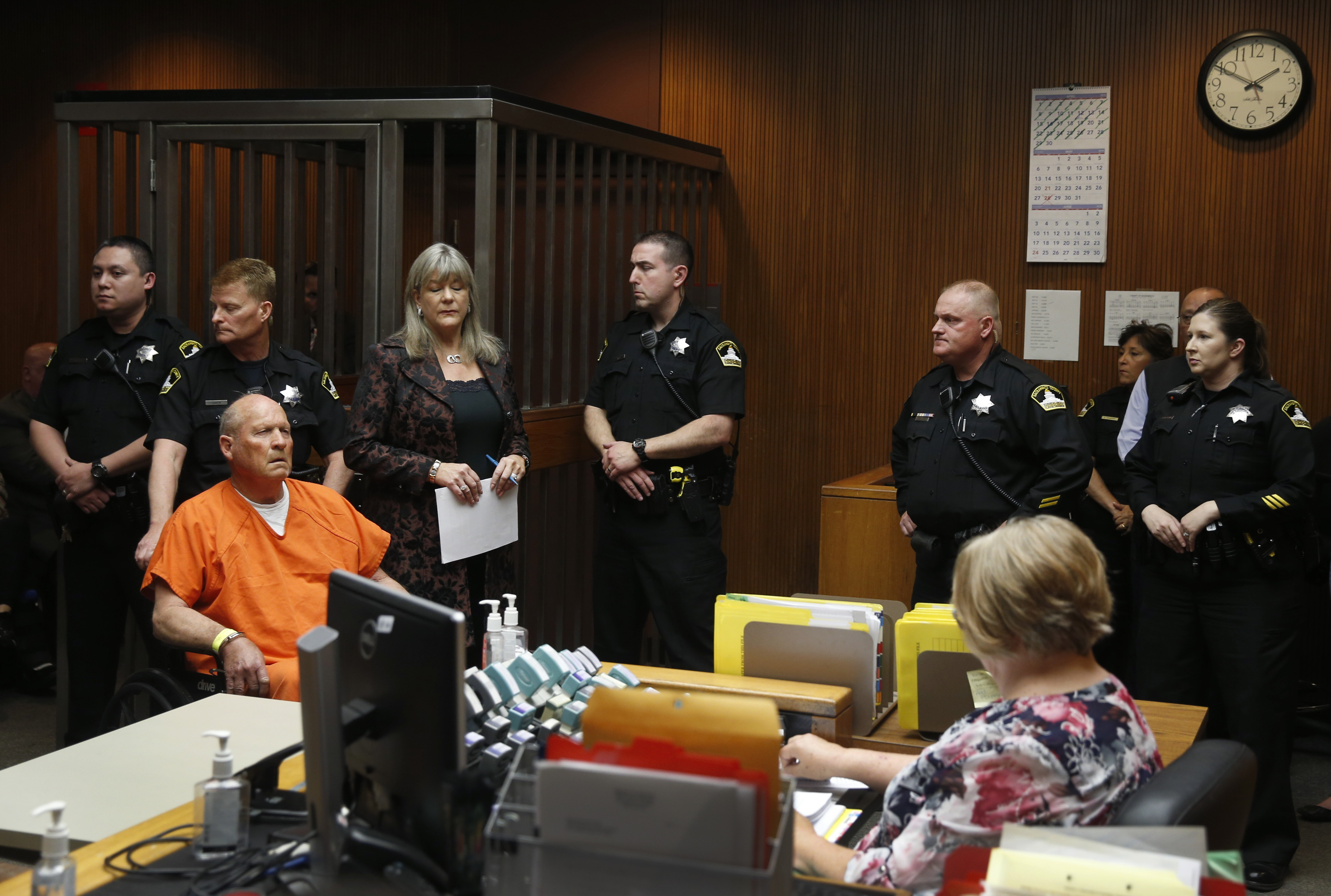 An elderly, balding Caucasian man in an orange prison uniform, seated in a wheelchair and surrounded by uniformed law enforcement officers in a courtroom. A woman with long graying hair stands to the man's left.