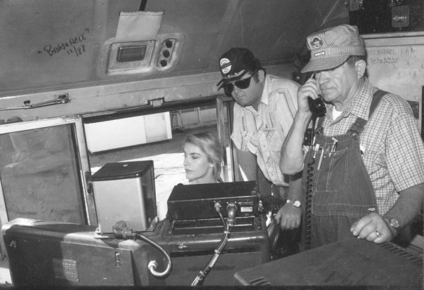 A woman sits at machinery in the conductor's cab of a train. Two men in caps stand beside her; one is holding a telephone receiver to his ear.