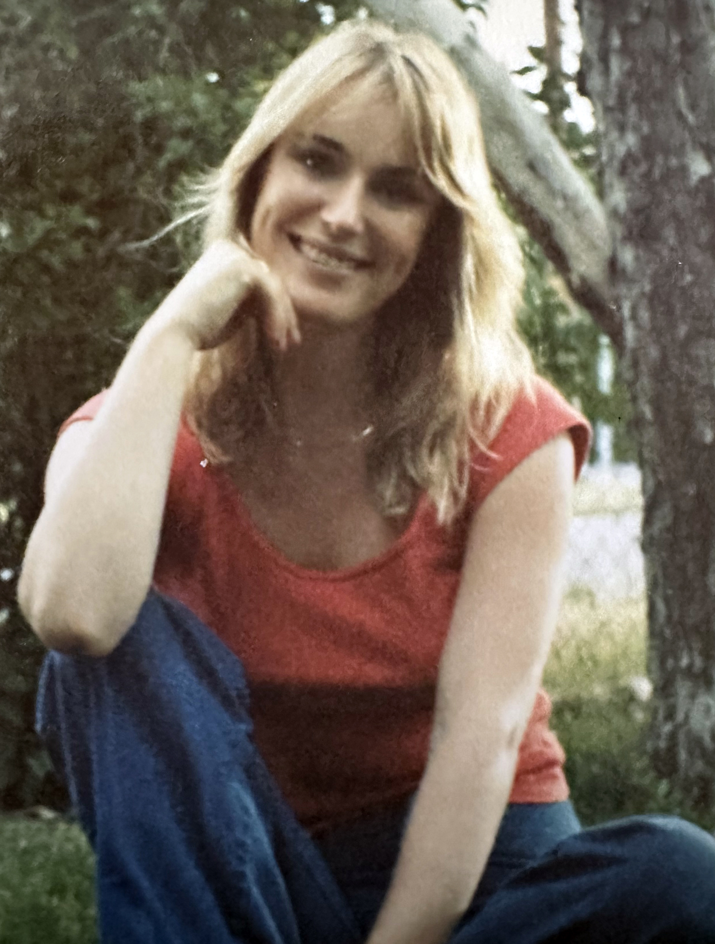 Blond woman wearing a short-sleeved red shirt and jeans, sitting outside, smiling.