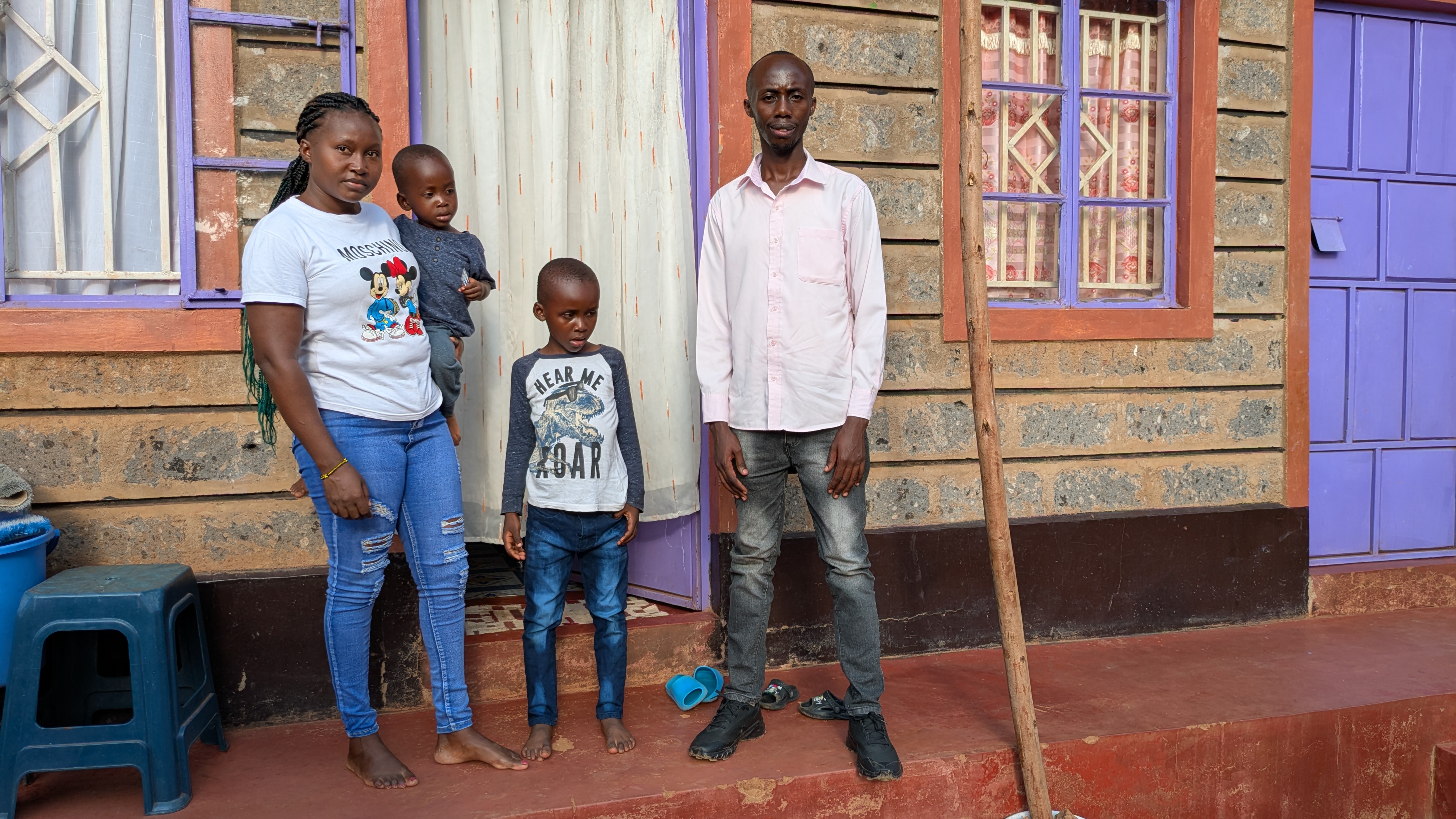 A young Kenyan family, including a husband, wife holding their toddler on her left hip, and their eldest child, stand in front of their home. The door and window frames are painted a vibrant lilac color.