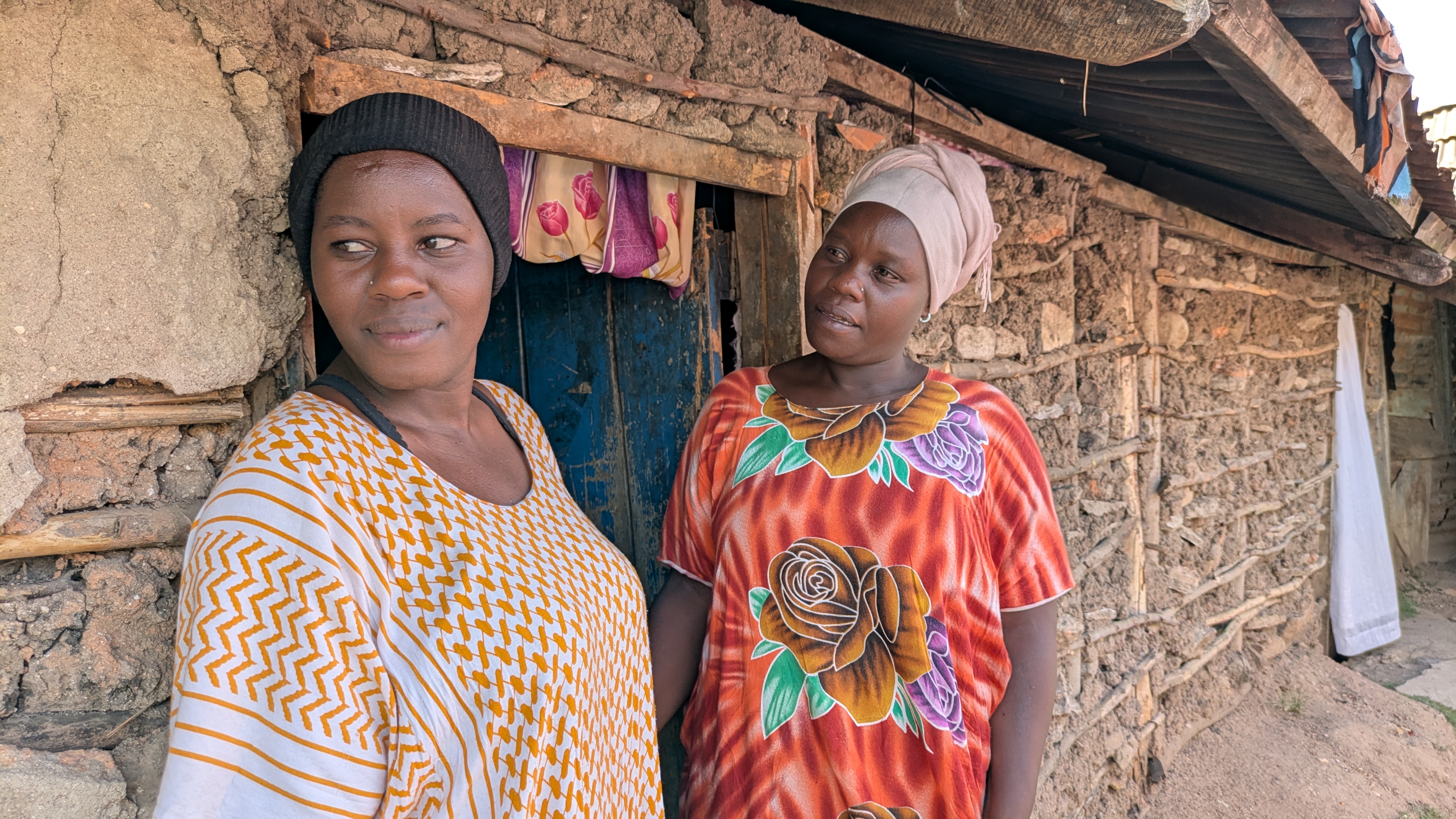 Two Kenyan women in boldly patterned tops stands outside their modest home, with walls made of packed earth, rocks, and wood.