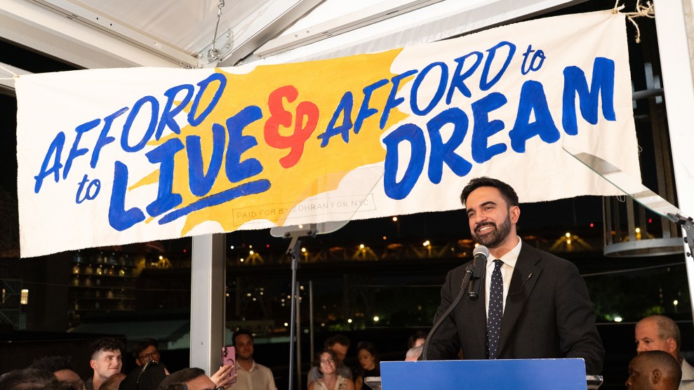 Zohran Mamdani wearing a black suit and tie stands in front of a banner that says "Afford to live and afford to dream."
