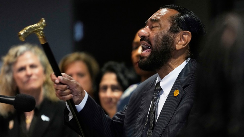 Texas Representative Al Green passionately speaks at a podium, holding a black cane with a gold handle in his raised hand. He is wearing a dark pinstripe suit and a patterned tie, and appears emotionally engaged. Several blurred individuals, mostly women, stand behind him in support.