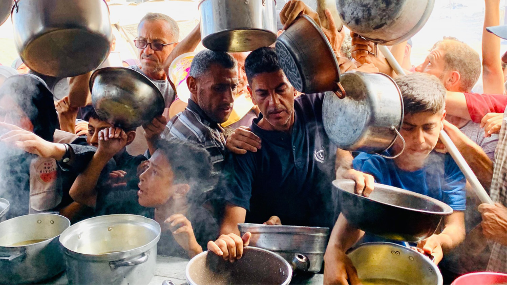 A crowd of men and boys hoist pots in anticipation of having them filled with food. Steam rises from a few partially filled pots on a counter in front of them.