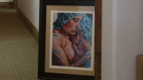 A photograph of a painting that's leaning against the wall as it stands on the floor. The painting shows a mother in a hospital bed holding a newborn child.