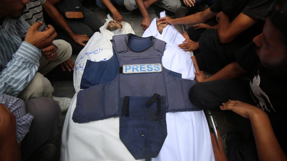 Mourners gather around two bodies of journalists killed Sunday in Gaza. The bodies are wrapped in white sheets and a press vest lays on top of them.