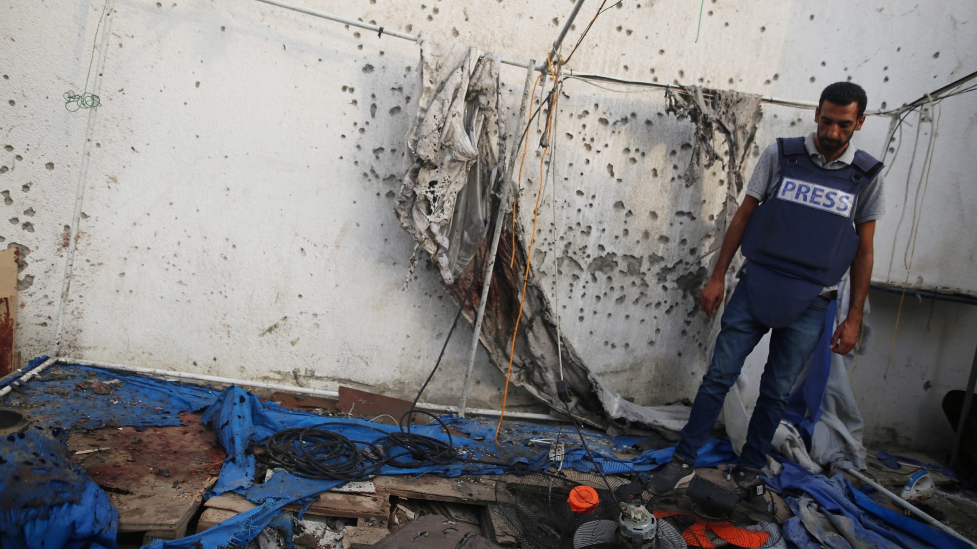 A man in a press vest stands inside a tent ridden with bullet holes.