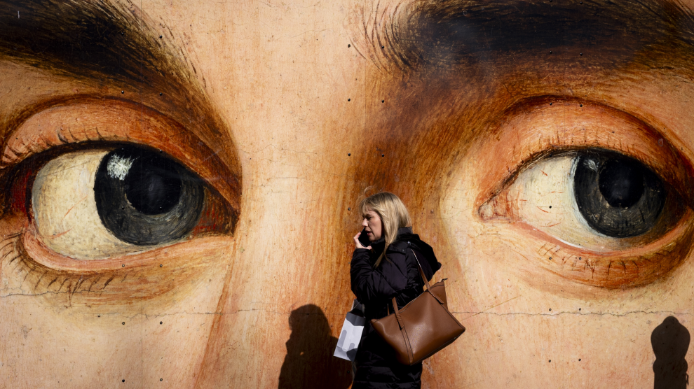 A Caucasian woman with hair down to her shoulders, wearing a coat and carrying two bags, walks to our left as she talks on a mobile phone. Behind her is a large mural of a face, seen from the bridge of the nose to the forehead. The dark gray eyes of the painting seem to be in line with the passing woman, whose shadow falls against the lower part of the wall.