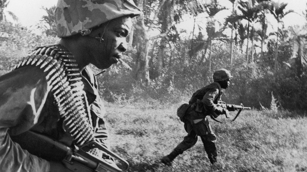 Two African American soldiers walk through a field lined with palm trees, carrying rifles.