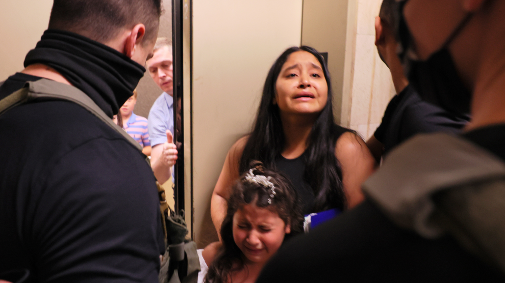 A young woman and girl, both with long dark hair, stand together crying in front of the partially open doors of an elevator. Standing across from them are masked agents wearing dark colors and tactical vests.