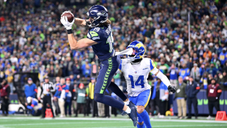 A Seattle Seahawks player makes a leaping catch for the football in the end zone in front of a trailing defender. A colorful crowd, out of focus, can be seen in the background.