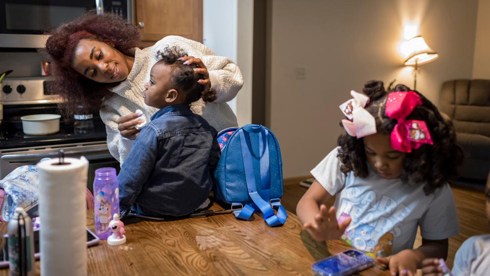A Black woman wearing a white sweater wipes the face of her young daughter while she sits on top of a kitchen table. Her eldest daughter, wearing pink hair ribbons and a blue Care Bears T-shirt, stands next to her, looking at a phone on the table.