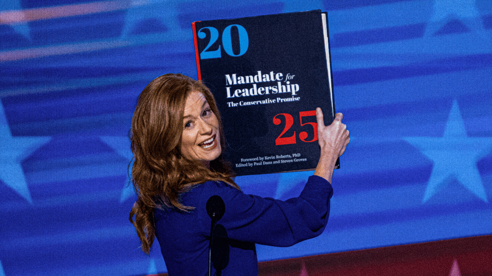 A middle-aged woman with long reddish-brown hair, dressed in a blue blouse, looks over her right shoulder as she speaks into a microphone on a stage. She holds up an oversized book with the title, “2025 Mandate for Leadership: The Conservative Promise.”
