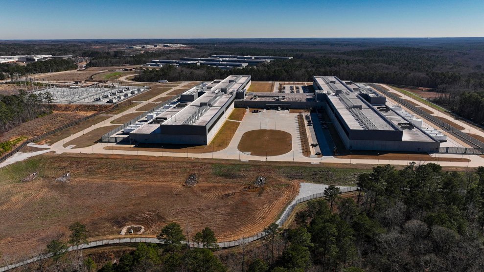 A drones-eye view of a large industrial comples consisting mostly of two long, flat, white buildings on a barren property, but with forest surrounding it. Sunny day.