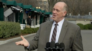 Tom Homan speaks to reporters (who are offscreen). He is wearing a gray suit. He is holding out his right arm while he speaks into several microphones.