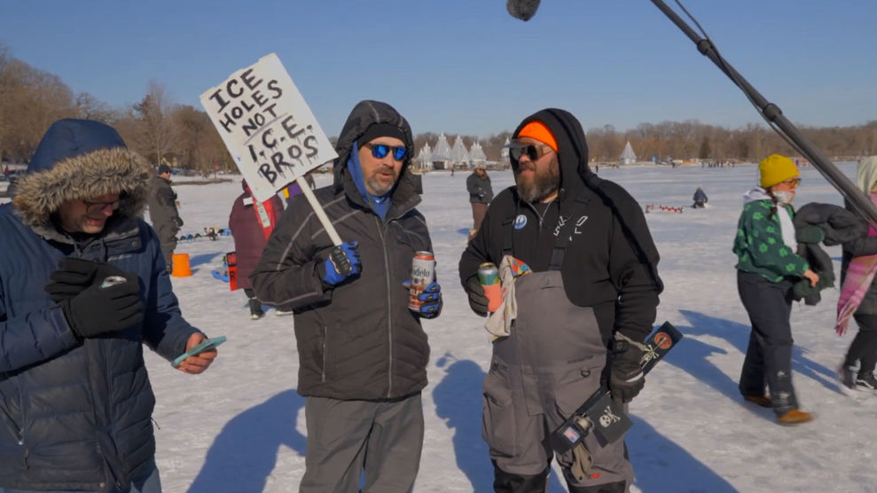 Two men dressed in winter outdoor clothing stand on a frozen lake, among a group of other people. One man holds a sign that reads, "Ice holes, not ICE bros."